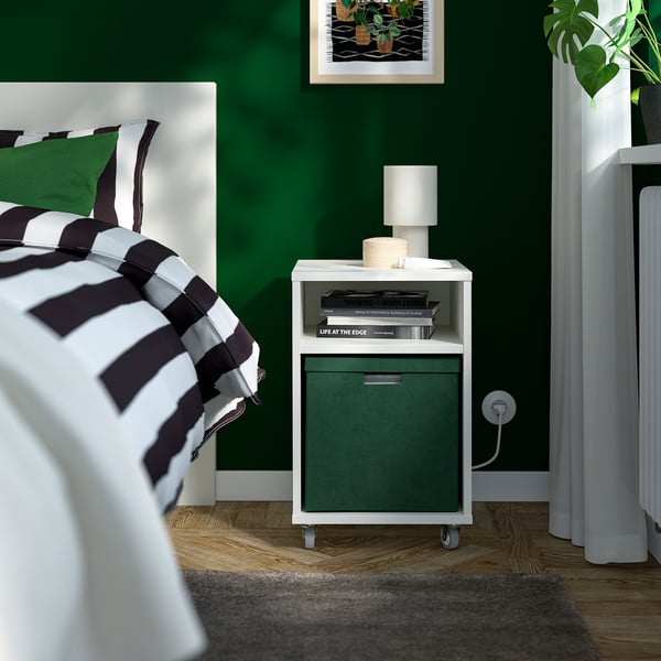 Green velvet GJÄTTA storage box with wheels, drawer, and shelf of books near bed.