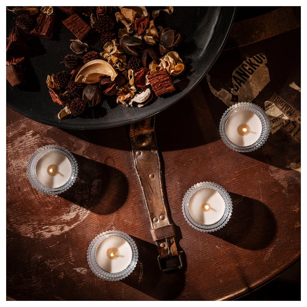 Four lit silver candles form a square around a black bowl filled with various dried plants on a worn wooden table. Soft glow illuminates the scene.