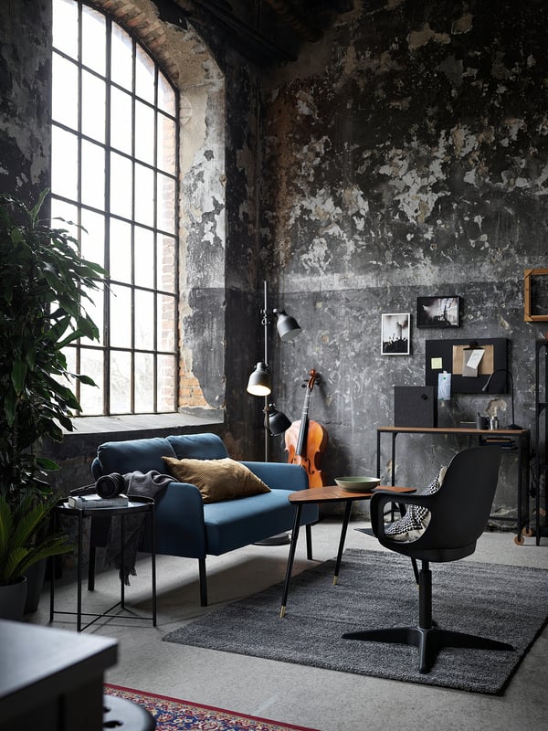 Industrial style living room with blue GLOSTAD sofa, modern desk, black chair, large window, plants, and rustic walls.