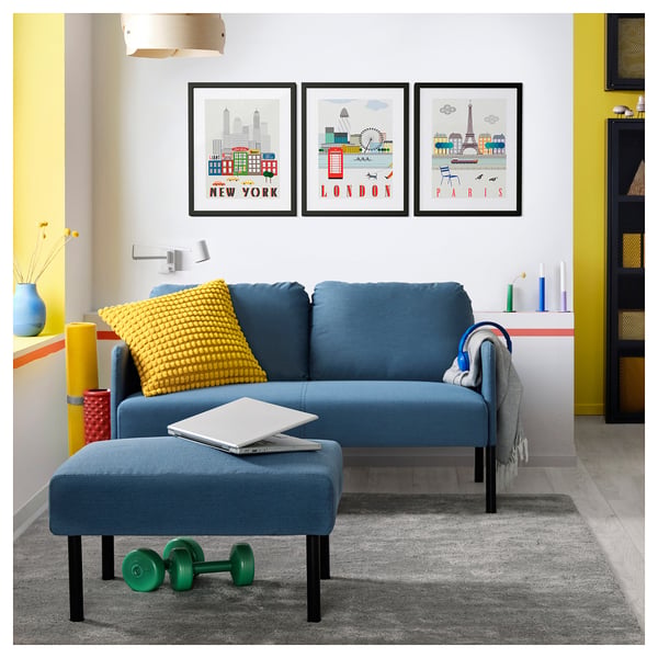 Modern living room with blue GLOSTAD sofa, yellow pillow, laptop, green dumbbells, grey rug, and city prints on wall.