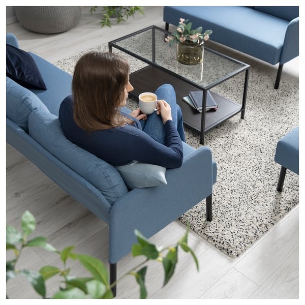 A person relaxes on a blue GLOSTAD sofa, holding a mug, set on a speckled rug with a black coffee table holding a vase of flowers.
