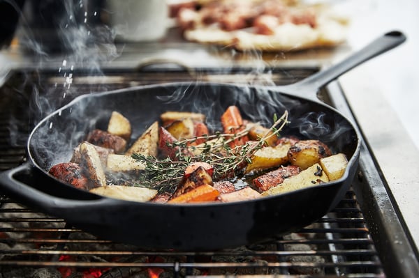 Black cast-iron skillet grilling colourful vegetables and herbs on a modern black grill with smoke rising.