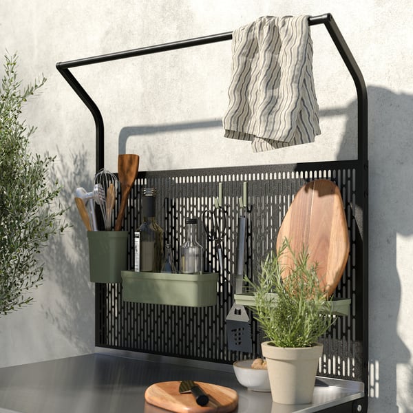 Modern kitchen rack with black metal grid, green shelf, hanging towels, wooden utensils, and potted plants.