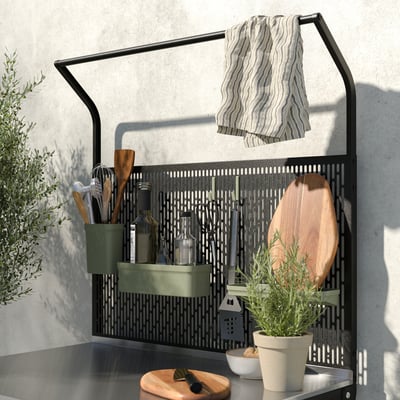 Modern kitchen rack with black metal grid, green shelf, hanging towels, wooden utensils, and potted plants.
