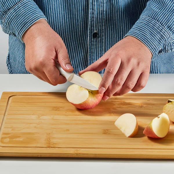 Person slices apple on wooden cutting board using small ceramic knife, demonstrating precise kitchen cutting.
