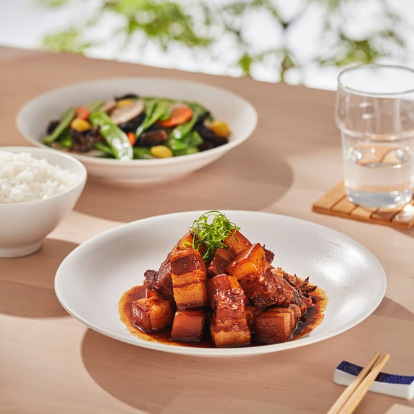 Plates of asian cuisine on wooden table. One with rice, another with vegetables, and one with braised meat, chopsticks nearby.