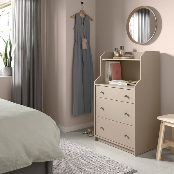 A HAUGA chest of drawers in beige with a round mirror above, placed in a pink room with a bed and plant.