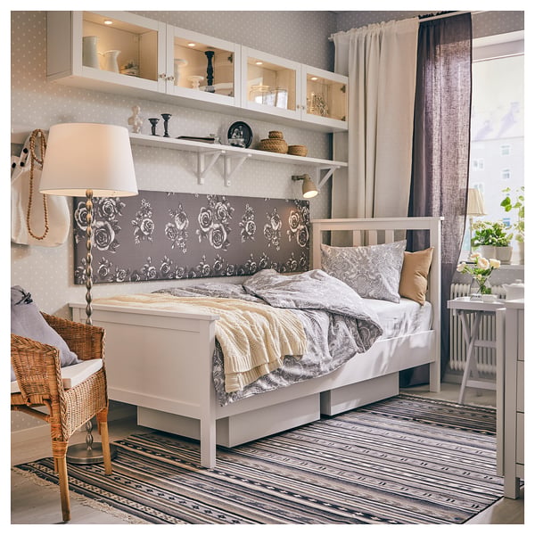 Cosy bedroom with white HEMNES bed, floral headboard, wicker chair, striped rug, and wall shelves.
