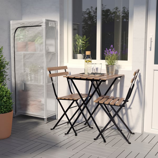 Small outdoor patio set with wooden table and two chairs, near a white wall with a window and a potted plant.