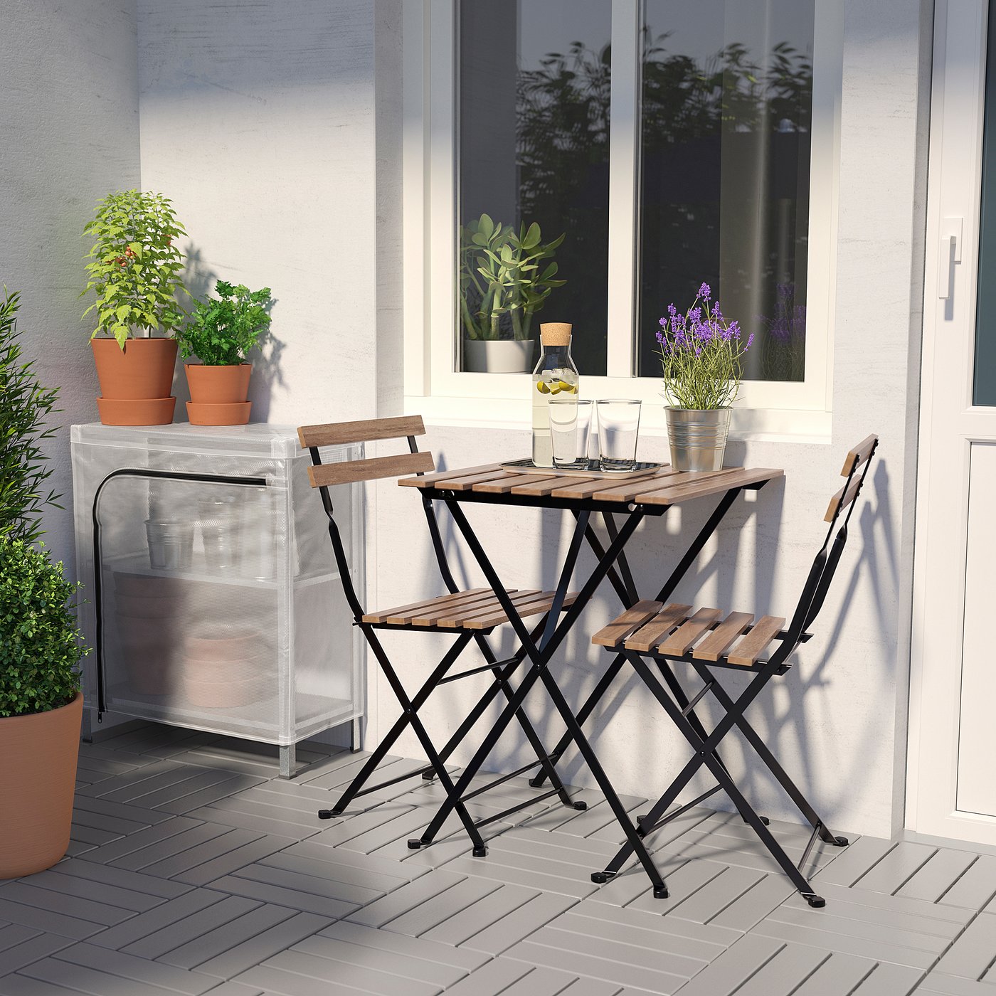 Cosy patio with small HYLLIS table and chairs. Surrounded by potted plants, placed near a white wall with windows.
