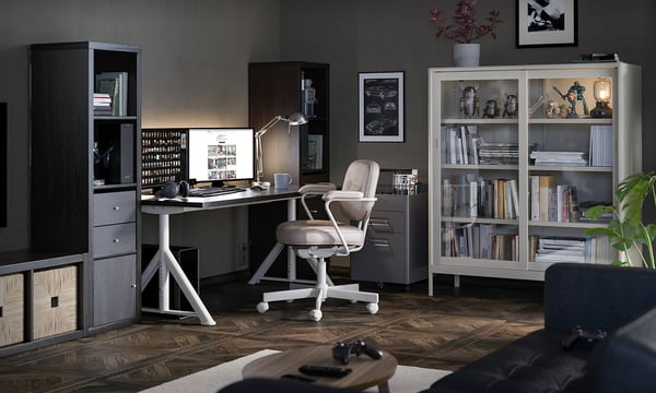 Modern home office with IDÅSEN desk, chair, and shelves. Desk is white, chair is grey, shelves hold books. Dark walls, wood floor.