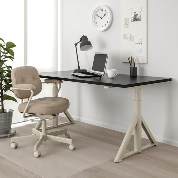 Modern home office with black adjustable desk, beige chair, clock, and plant.