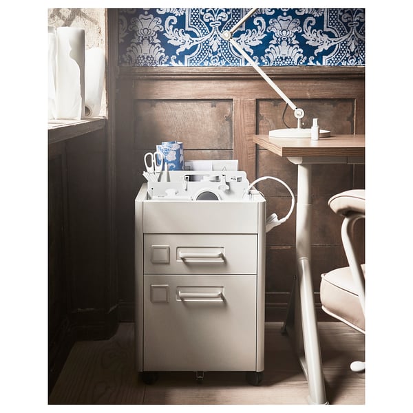 An IDÅSEN silver metal filing cabinet with three drawers, standing on wheels, in a room with patterned wallpaper.