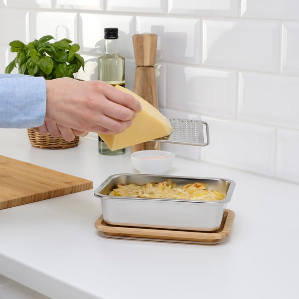 Person grates cheese over baked dish in stainless steel food container with bamboo lid on counter.
