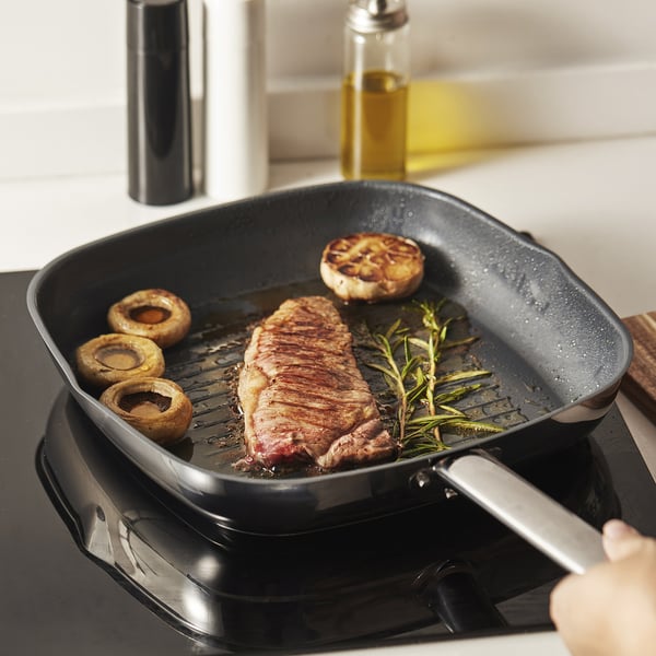 A grill pan with steak, mushrooms, and herbs cooking, showcasing non-stick surface and even heat distribution.