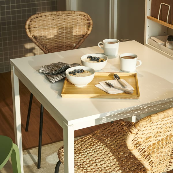 Square white table set with bowls of cereal, coffee cups, napkins, and spoons. Surrounded by woven chairs.