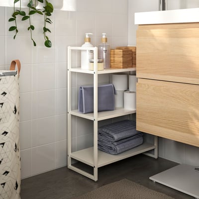 White JONAXEL storage unit in bathroom. 3-tiered, holds towels, liquids, and boxes. Fits neatly beside wooden vanity.