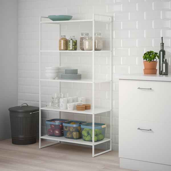 JONAXEL white metal shelf unit holds kitchen items, including jars, bowls, plates, and fruits in baskets, fitting neatly beside counter and cabinet.