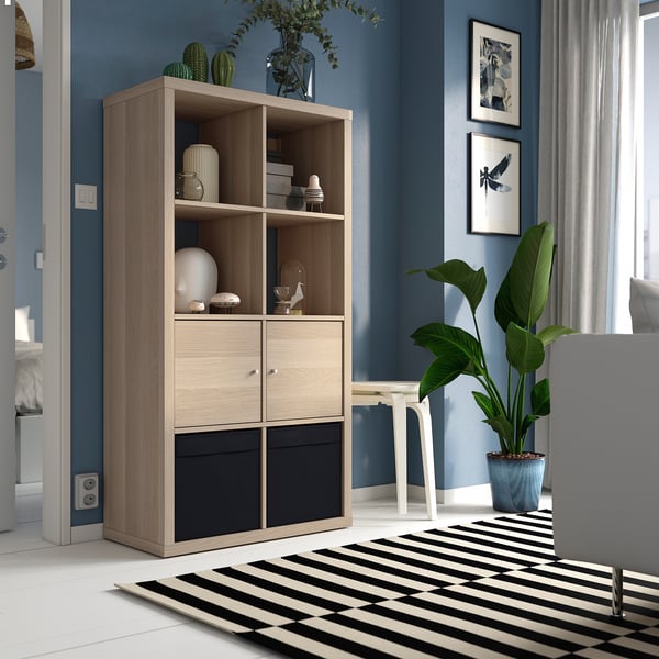 KALLAX light wood shelf with drawers in blue room, holding vases, books, and décor. Surrounded by rug, plant, and chair.