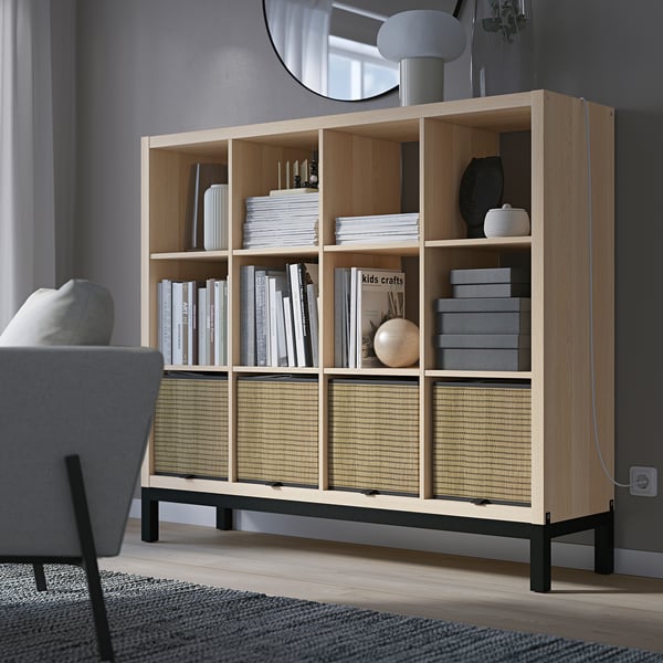 KALLAX shelving unit with books, decor, and wicker inserts in a room. Grey sofa and lamp visible.