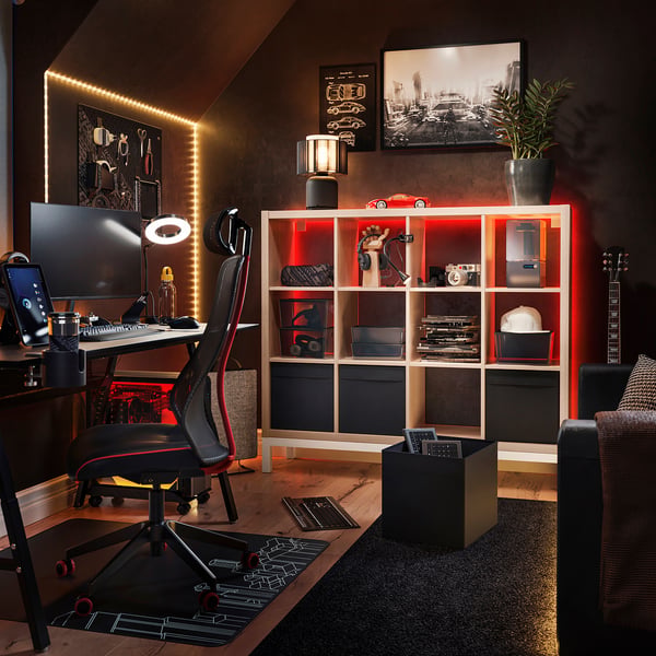 Modern home office with black KALLAX shelf unit, red led lights, black desk, black computer chair, monitor, keyboard, and guitar.