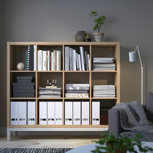 KALLAX shelving unit stores books, games, and decor. Tall, light wood, rectangular, with multiple compartments. Fits well in living spaces.