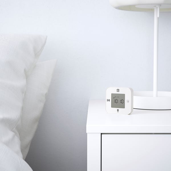 A modern bedside alarm clock displaying time on a white bedside table with a lamp and crisp white bedding.