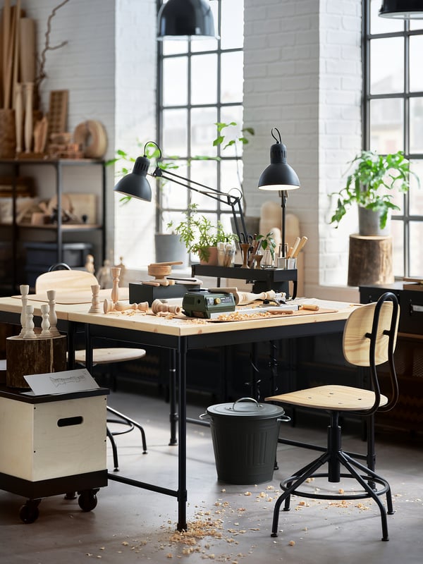 Industrial-style workspace featuring KULLABERG black metal desk with wooden top, surrounded by tools, plants, and black chairs.