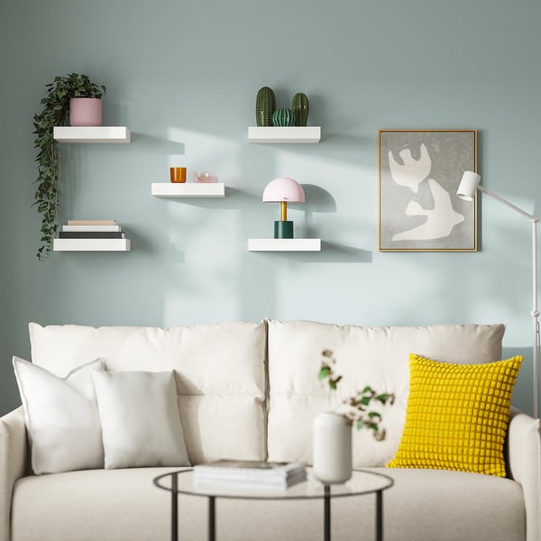 Modern living room with blue walls, white couch, colourful pillows, & minimalist white LACK shelves displaying books, vases, plants, and a lamp.