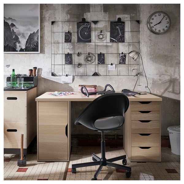 Modern office setup with LAGKAPTEN light brown desk, black swivel chair, wall clock, and decorative wire grid with photos.