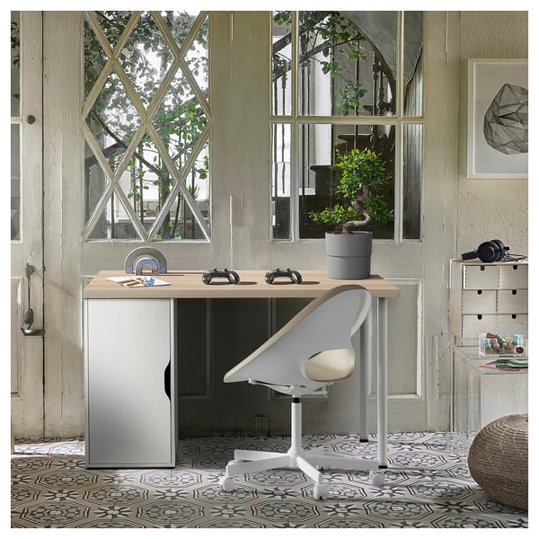 Home office with wooden desk, grey cabinet, white chair, large window, plant, office supplies, geometric print on textured wall, patterned rug.