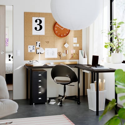 Home office with black LAGKAPTEN desk, chair, computer, and supplies. Large corkboard and clock above, window and plant beside.