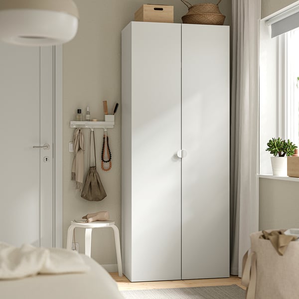 A platsa LASTARE white wardrobe near a window, taller than a nearby chair, with hanging items and decorations on top.