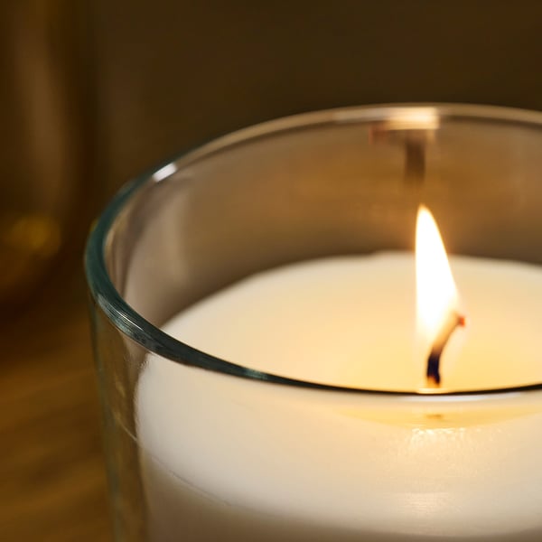Burning scented candle in clear glass, emitting soft light, with a calm flame and creamy wax inside.