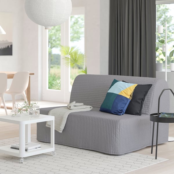 Modern, grey sofa-bed with colourful pillows. Sleek, rectangular design fits well in living spaces, matching white tables and large windows.