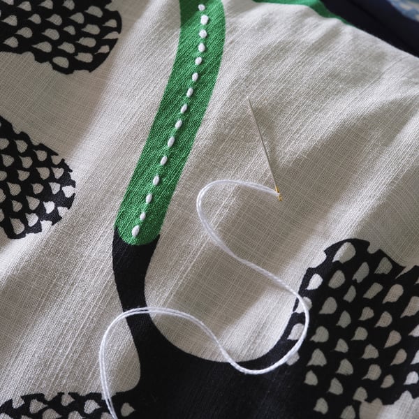 Needle with thread sewing white fabric with green, black, and white pattern; detailed handiwork.