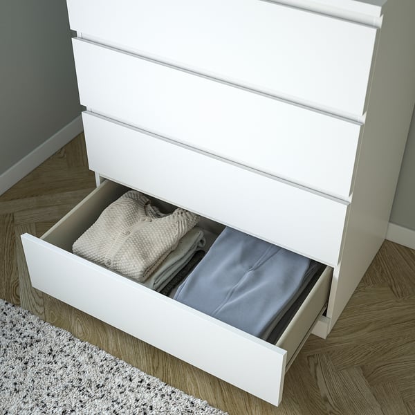 Open white MALM dresser with folded clothes inside. Drawers are smooth and can store multiple items. Safety feature noted as wall-anchoring.