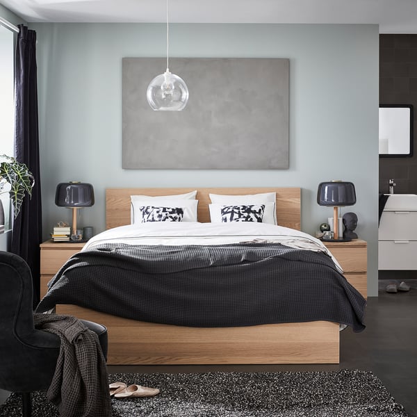 Modern bedroom with light wood bedframe, grey bedding, black lamps, and abstract art.