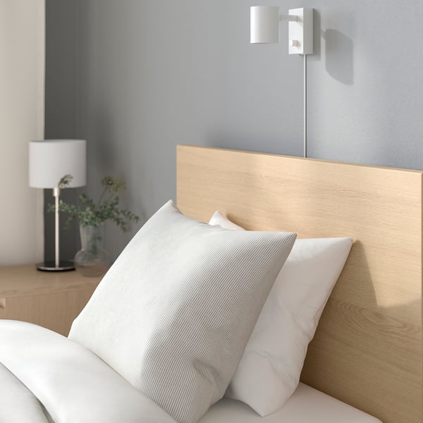 Modern bedroom with light wooden headboard, white bedding, minimalist lamp, and decorative plant.