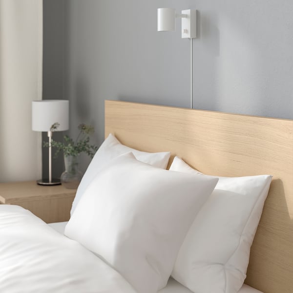 A minimalist, wooden bed with white pillows and a bedside lamp. A wall-mounted light switch is visible above the bed.