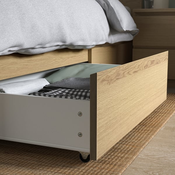 Open wooden bed drawer revealing folded clothes and linens on wheels.