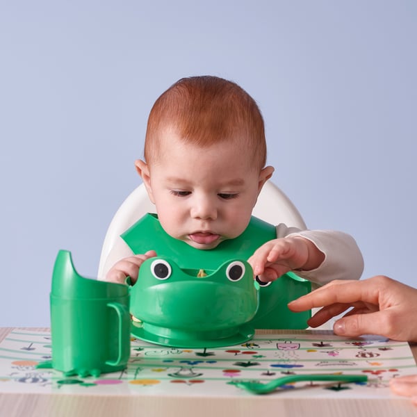 Baby sits in high chair, holding green frog-shaped cup with wide handles. Adults hand holds spoon near colourful mat. Frog bib catches spills.