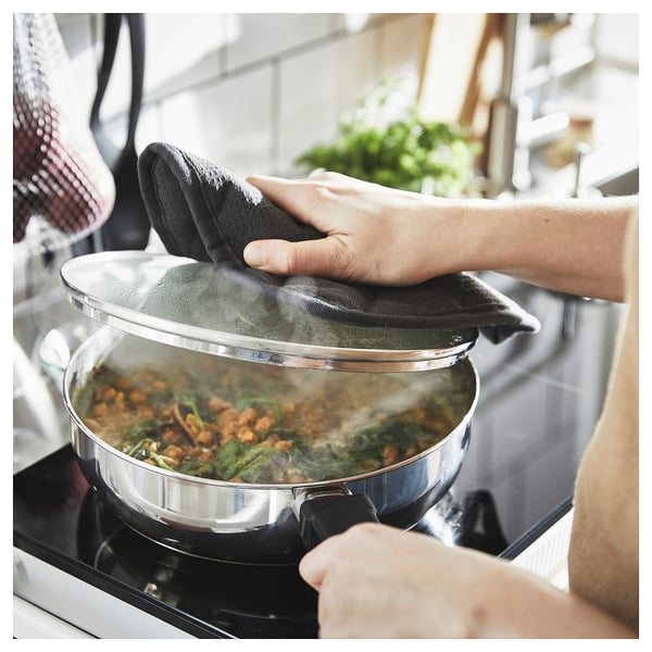 Person cooking vegetables in a stainless steel frying pan with a transparent lid on a stove.