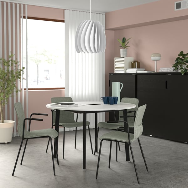 Modern round white MITTZON conference table with green chairs by window. Black cabinet with books and a blue mug. Small, ideal for small groups.