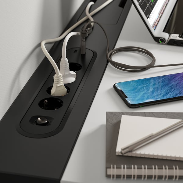 Black wireless charging pad on desk with plugged-in devices and notebook.