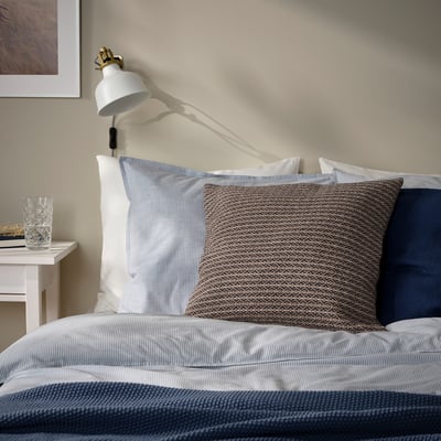 Bed with MOROTSMOTT grey-beige cushion, coarsely-textured, wall lamp.