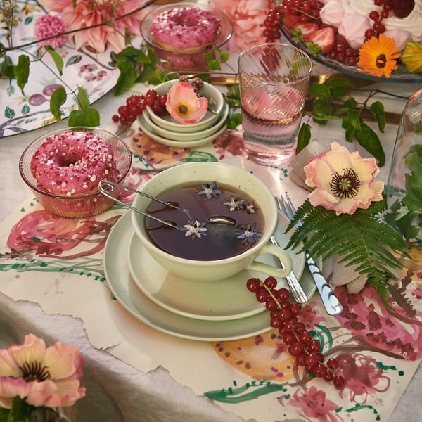 Elegant table set with green mug, glass of water, pink pastries, flowers, and fern leaves on floral tablecloth.