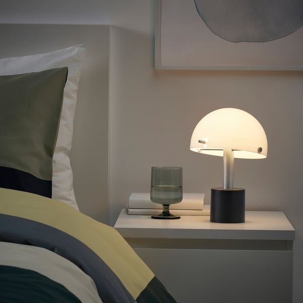 Small, stylish lamp on bedside table with soft light, grey base, white shade, green vase, book. Cosy room with striped bedding.