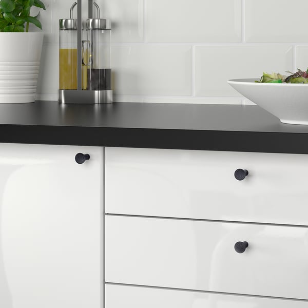 Modern kitchen with white cabinetry and black handles; a bowl of salad sits on a black countertop against a white brick backsplash.