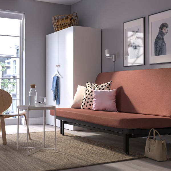 Modern living room with NYHAMN terracotta sofa-bed, low-profile, large, pink & polka-dot pillows. Side table with jug and glass, beige bag on rug.